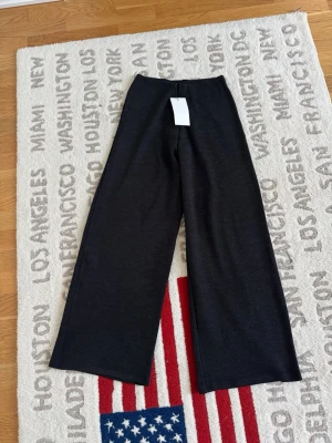 Vida svarta byxor i stickad look - Snygga vida byxor i svart med stickad känsla och hög midja. Materialet är mjukt och stretchigt, perfekt för en avslappnad och trendig stil. Passar till oversized tröjor eller crop tops för en cool vardagslook.