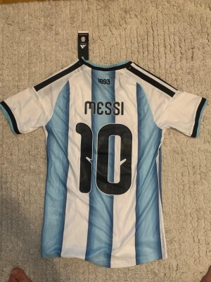 Argentina Messi matchtröja Adidas - Säljer en officiell Argentina matchtröja från Adidas med Messi och nummer 10 på ryggen. Tröjan har klassiska blå och vita ränder, svarta detaljer och guldiga logotyper framtill. Tillverkad i lätt och ventilerande funktionsmaterial, perfekt för fotboll.