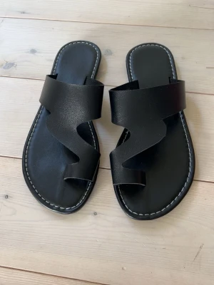 Svarta slip-in sandaler i läderlook - Säljer ett par stilrena svarta slip-in sandaler med breda remmar i konstläder och vit kontrastsöm runt sulan. Sandalerna har en modern, vågig design på remmarna och en platt sula med räfflad undersida för bättre grepp. Perfekta för sommardagar och enkel att matcha.