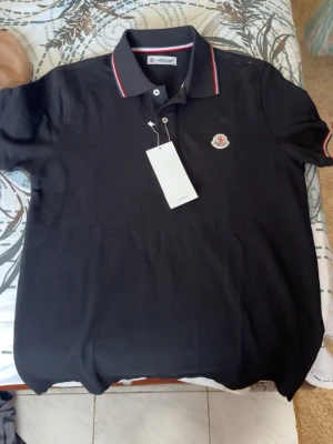 Svart pikétröja från Moncler - Svart pikétröja från Moncler med klassisk krage och knäppning framtill. Tröjan har röda och vita ränder på krage och ärmslut samt Moncler-logga på bröstet. Perfekt för en stilren och sportig look.