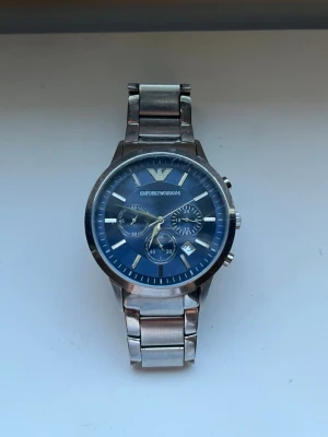 Emporio Armani blå klocka i stål - Stilren klocka från Emporio Armani med blå urtavla och silverfärgat armband i rostfritt stål. Klockan har flera små urtavlor, datumvisning och klassisk design med märkets logga högst upp. Perfekt accessoar för en modern look. Ny pris 2300