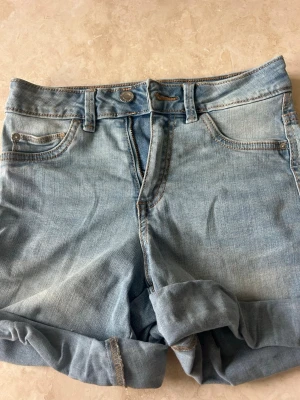 Ljusblå denimshorts med uppvik - Snygga ljusblå denimshorts med uppvikta benslut och klassisk femficksdesign. Shortsen har bälteshällor, knapp och dragkedja framtill. Perfekta för varma dagar och enkla att matcha med olika stilar.