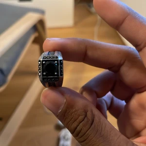 Silverfärgad ring med svart sten - Snygg silverfärgad ring med en stor svart fyrkantig sten i mitten och små svarta detaljer runt om. Ringen har ett coolt mönster längs sidorna och känns riktigt stilren och unik. Perfekt för dig som vill sticka ut med dina accessoarer.