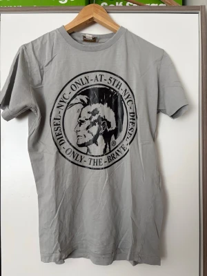 Grå vintage t-shirt från Diesel  - Snygg grå t-shirt från Diesel med stort svart tryck framtill och rund halsringning. T-shirten har korta ärmar och är tillverkad i mjuk bomull, perfekt för en avslappnad streetstyle-look.