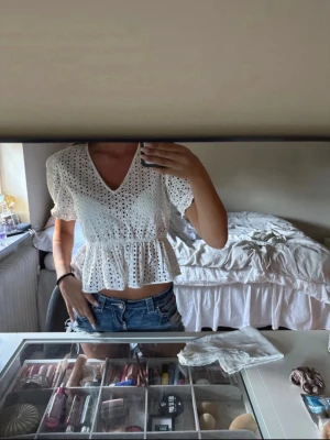 Vit spetsblus med volang och v-ringning - Söt vit blus i bomull med broderad spets, v-ringad hals och volang nedtill. Toppen har korta puffärmar och är croppad, perfekt till jeans eller kjol. Luftig och somrig känsla med fina detaljer.