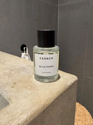 ESSNCE Belle Femme Eau de Parfum - En stilren parfym från ESSNCE med namnet Belle Femme. Flaskan är cylindrisk i genomskinligt glas med svart rund kork och enkel vit etikett. Doften är en Eau de Parfum och flaskan har en modern, minimalistisk design som passar perfekt på badrumshyllan.