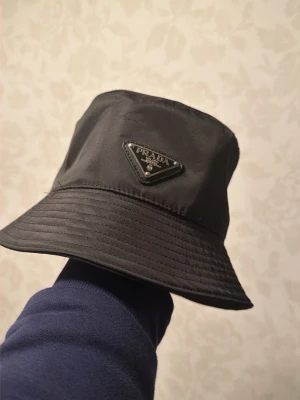 Svart bucket hat från Prada - Har en bucket hat från Prada jag inte använt för mycket den passar skönt och är perfekt inför de varma sommar vädret. (Står no size men är storlek M)