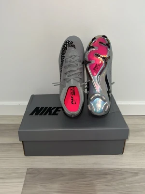 Nike grå Mercurial fotbollsskor - Snygga grå Nike Mercurial fotbollsskor med svart swoosh och rosa detaljer på innersulan och yttersulan. Skorna har en modern, strömlinjeformad design och en glansig, mönstrad yttersula för bästa grepp på planen. Perfekta för dig som vill sticka ut på matchen.
