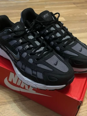 Nike p6000 - Nike p6000 sneakers i svart och grått med mesh- och syntetpaneler, svarta snören och vit mellansula. Swoosh-logga på sidan och detaljerad design som ger en sportig vibe. Perfekta för dig som gillar streetwear och snygga detaljer.
