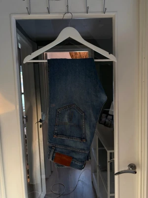 Blå raka jeans från tiger of sweden - Snygga blå jeans med rak passform och klassisk femficksdesign. Jeansen har en unik bakficka med dekorativa sömmar och en brun läderpatch vid linningen. Perfekta till en avslappnad streetstil. OBS, dessa har hål i bakfickan.