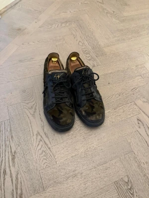 Kamouflage sneakers från Giuseppe Zanotti - Snygga sneakers i kamouflagemönster från Giuseppe Zanotti. Skorna har svarta detaljer, dragkedjor på sidorna och klassiska svarta snören. Yttermaterialet är en mix av mocka och läder, med en svart gummisula. Perfekta för dig som vill sticka ut med en cool streetstyle. Nyrpis 6500