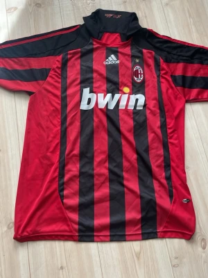 AC Milan Ronaldinho matchtröja - Säljer en klassisk AC Milan matchtröja från Adidas med Ronaldinho och nummer 80 på ryggen. Tröjan är röd och svart med vertikala ränder, klubbmärke på bröstet och Bwin som sponsor. Perfekt för dig som älskar fotboll och vill ha en ikonisk supportertröja.