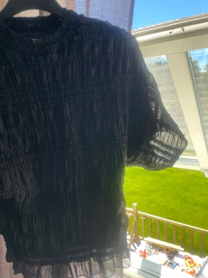 Svart meshblus från Nelly - Trendig svart meshblus från Nelly med korta ärmar och rynkade detaljer över hela tyget. Blusen har en rund halsringning och är tillverkad i ett tunt, transparent material som ger en luftig känsla. Perfekt att styla lager på lager.