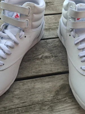Vita Reebok Freestyle Hi sneakers - Snygga vita Reebok Freestyle Hi sneakers med klassisk design. Skorna har högt skaft, dubbla kardborreband och vita skosnören. Tillverkade i slätt läder med diskret Reebok-logga på sidan och tungan. Perfekta för en clean och sportig look.