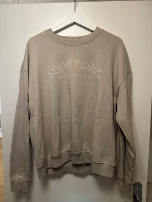 Beige sweatshirt med tryck - Beige sweatshirt från Lager 157 med texten 'NEW YORK UPPER EASTSIDE' tryckt på bröstet. Tröjan har rund hals, långa ärmar och ribbade muddar. Mjuk insida och avslappnad passform, perfekt för en chill look.