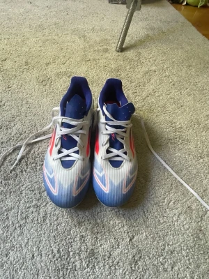 Adidas F50 vita/blå/röda fotbollsskor - Säljer ett par Adidas F50 fotbollsskor i vitt med blå och röda detaljer. Skorna har klassisk snörning, låg profil och dobbar för spel på gräs. Snygg design med tydliga Adidas-ränder på sidan och en sportig look som sticker ut på planen.frakt betalas av köpare