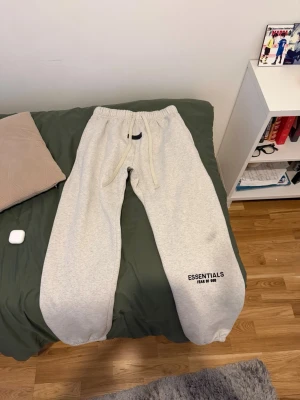 Grey Essentials Fear of God pants/joggers - Snygga ljusgrå mjukisbyxor från Fear of God Essentials med svart logga på benet. Byxorna har resår i midjan med dragsko och muddar vid bensluten. Tillverkade i mjukt och bekvämt material, perfekta för chill dagar.