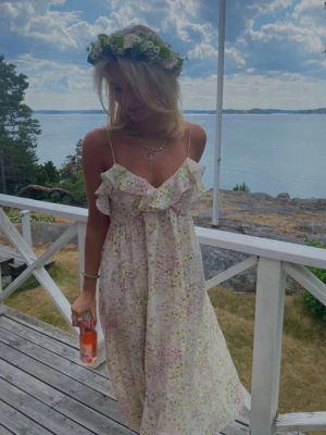 Blommig långklänning med volang - Romantisk långklänning med smala axelband och volangdetalj vid bysten. Klänningen har ett blommigt mönster i ljusa pastellfärger på vit botten, perfekt för sommarens tillfällen. Luftigt material och feminin siluett.