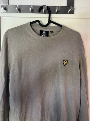 Beige stickad tröja Lyle & Scott - Snygg beige stickad tröja från Lyle & Scott i storlek M med rund halsringning och den klassiska gula örnloggan broderad på bröstet. Tröjan har långa ärmar och är tillverkad i stickad material som passar perfekt till vardagslooken. Skriv vid funderingar!