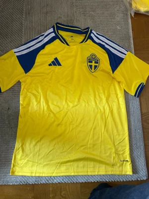 Svensk landslagströja Adidas gul/blå - Gul och blå fotbollströja från Adidas med Sveriges landslagsemblem på bröstet. Tröjan har blå detaljer på axlar och ärmar, samt vita ränder längs axlarna. Tillverkad i lätt och ventilerande material med Climalite-teknologi.