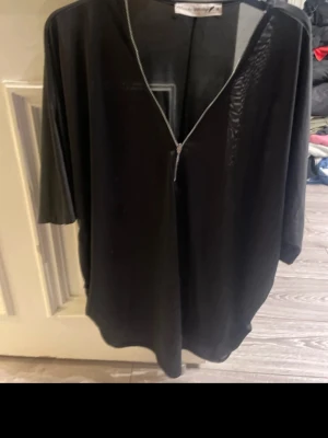 Svart omlottopp med dragkedja - Snygg svart omlottopp från Madeleine Vionnet med dragkedja framtill. Toppen har trekvartsärm och en lös, oversized passform. Materialet känns lätt och luftigt, perfekt för att styla med olika accessoarer.