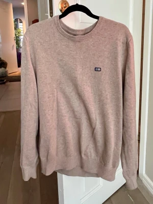 Beige tröja från Lexington - Snygg beige tröja från Lexington med rund halsringning och diskret logga på bröstet. Tröjan har långa ärmar och är tillverkad i mjukt, tunt stickat material som känns skönt mot huden. Perfekt för en clean och stilren look.