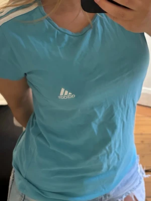Blå Adidas t-shirt med vit logga - Snygg blå t-shirt från Adidas med vit logga på bröstet och vit rand på axeln. Klassisk passform och rund halsringning. Perfekt till en avslappnad stil och enkel att matcha med jeans eller shorts.