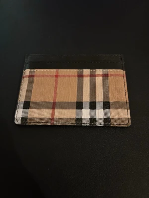  Kortfodral - Snyggt kortfodral i klassiskt beige, svart, vitt och rött rutmönster. Tillverkat i tåligt material med svarta detaljer och flera kortfack. Perfekt för dig som vill ha en stilren och ikonisk accessoar i fickan.