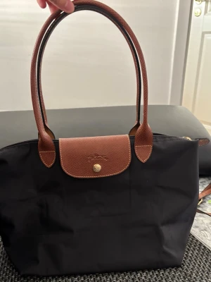 Svart Longchamp Le Pliage handväska - Klassisk svart handväska från Longchamp Le Pliage med bruna läderhandtag och lock. Väskan är tillverkad i slitstark nylon med detaljer i brunt läder och har guldiga metalldetaljer. Perfekt storlek för att bära det viktigaste och enkel att matcha till olika outfits.