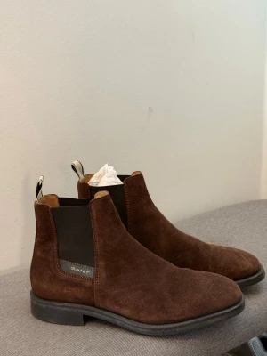 Bruna Chelsea boots från Gant - Snygga Chelsea boots i mörkbrunt mocka från Gant. Klassisk siluett med elastiska paneler på sidorna och dragflikar bak. Skorna har en låg klack och svart sula, perfekta för en stilren look.