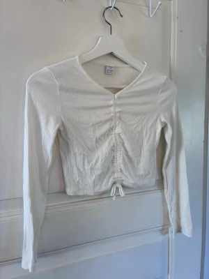 Vit ribbad topp för flickor från Lindex - Superfin vit långärmad topp för flickor från Lindex med ribbad struktur och snörning framtill. Toppen har v-ringning och är croppad, vilket ger en trendig look. Perfekt att styla med jeans eller kjol för en fräsch outfit.