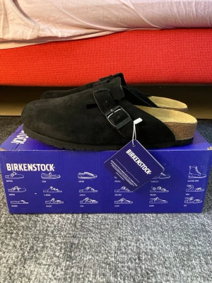 Svarta Birkenstock Boston tofflor - Svarta Birkenstock Boston tofflor i mocka med justerbart spänne och klassisk korksula. Snygg och tidlös modell som är superbekväm och passar perfekt för chill dagar hemma eller när du vill ha en avslappnad stil.