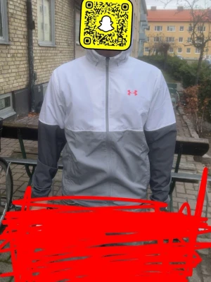 Under Armour gråvit vindjacka  - Snygg vindjacka från Under Armour i tvåfärgad design med vit ovandel och ljusgrå nederdel och ärmar. Jackan har dragkedja framtill, huva och elastiska muddar. Under Armour-logga i rött på bröstet. Perfekt för blåsiga dagar och sportiga outfits.
