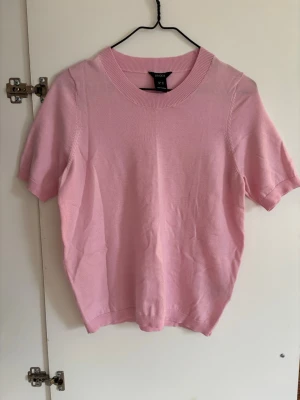 Ljusrosa stickad topp från Lindex  - Supersöt ljusrosa stickad topp från Lindex med korta ärmar och rund halsringning. Perfekt att styla med jeans eller kjol för en fräsch look. Mjuk och skön kvalitet som känns lyxig mot huden.