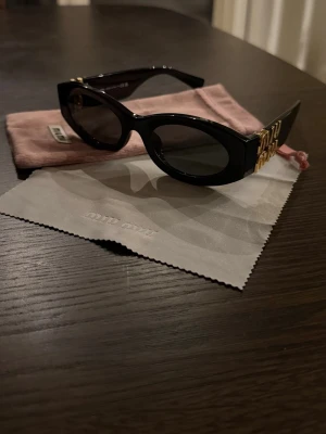 Svarta solglasögon från Miu Miu - Snygga svarta solglasögon från Miu Miu med ovala bågar och guldiga detaljer på sidorna. Glasögonen har en trendig och chunky design, perfekt för att lyfta din stil. Kommer med originalfodral och putsduk. Använd 1 sommar, men i fint skick 