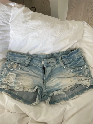 Ljusblå jeansshorts med spetsdetaljer - Säljer så snygga ljusblå jeansshorts i nyskick med slitna detaljer, nitar vid fickorna och spets på bakfickorna. Shortsen är lågmidjade.  Midiemåttet tvärs över är ca 36 och passar XS, längd 21cm💞säljs då de är för små.