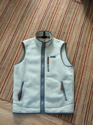 Vit fleeceväst från Patagonia - Snygg vit fleeceväst från Patagonia i storlek M. Västen har grå detaljer, dragkedjor med läderdetaljer och hög krage. Perfekt för lager-på-lager och outdoor-stil. Klassisk Patagonia-logga på bröstet och två fickor med dragkedja.
