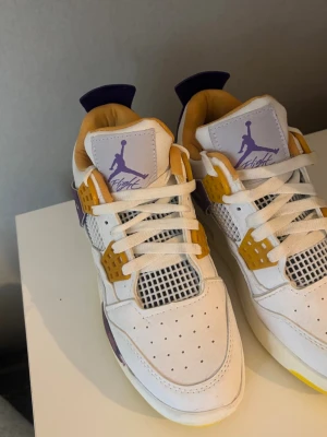 Air Jordan 4 Retro Lakers - Snygga Air Jordan 4 Retro sneakers i vitt läder med lila och gula detaljer. Klassisk mesh på sidorna och tungan, samt ikonisk Jumpman-logga i lila på tungan. Skön vadderad insida i gult och synlig Air-sula. 