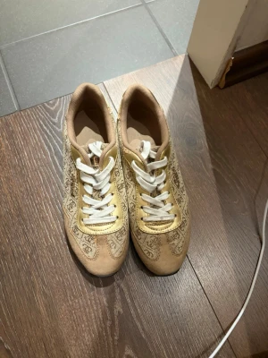 Beige sneakers med mönster och vita snören - Snygga beige sneakers med gulddetaljer och unikt mönster längs sidorna. Skorna har vita snören och en svart räfflad gummisula. Perfekta för dig som vill sticka ut med en cool och annorlunda stil. Materialet är en mix av textil och syntet.