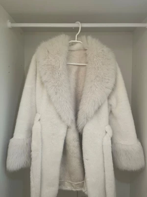 Vit fuskpäls kappa med bälte - Snygg vit kappa i fuskpäls med bred krage och stora muddar. Kappan har ett matchande bälte i midjan och två fickor framtill. Perfekt för dig som vill ha en lyxig och trendig look under kalla dagar.