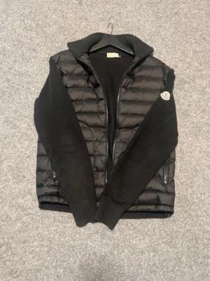 Moncler cardigan - Snygg svart pufferjacka från Moncler med stickade ärmar och hög krage. Jackan har dragkedja framtill, två fickor med dragkedja och Moncler-logga på ena ärmen. Perfekt för kalla vinterdagar och riktigt stilren look.