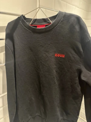 Svart sweatshirt från Hugo - Svart sweatshirt från Hugo med röd logotyp på bröstet. Klassisk rund halsringning och långa ärmar. Tröjan har en enkel och stilren design, perfekt för en avslappnad look. Materialet ser ut att vara mjukt bomullstyg.