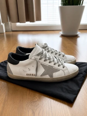 Golden Goose - Säljer ett par vita Golden Goose sneakers med grå mockadetaljer, grå stjärna på sidan och svarta hälpartier. Skorna har grå snören och GGDB-logga i guld på sidan. Klassisk låg modell med slitstark gummisula och cool vintagekänsla.