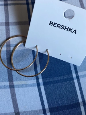 Guldfärgade hoops från Bershka - Stora, tunna örhängen i klassisk hoop-modell från Bershka. De är guldfärgade och har en enkel, stilren design utan extra detaljer. Perfekta för dig som gillar minimalistiska accessoarer.