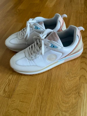 Vita sneakers från Palpa - Snygga vita sneakers från Palpa med detaljer i ljusrosa, beige och blått. Skorna har klassisk låg profil och en mix av läder,tyg och mocka i materialet. De är i gott skick.