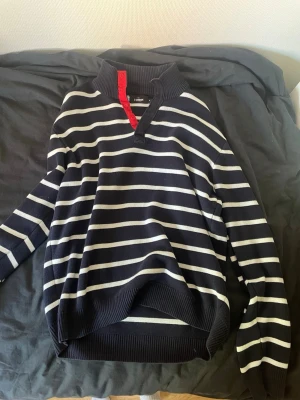 IVANHOE tröja - Tröja med långa ärmar och krage. Tröjan har ett randigt mönster i mörkblått och vitt. Kragen har en röd detalj med knappar. Sparsamt använd. Passar mig som är 183 ganska baggy, kan fixa mått vid fråga. Har en lite fläck på de vita, mitt på magen. Men den syns knappt vid användning.