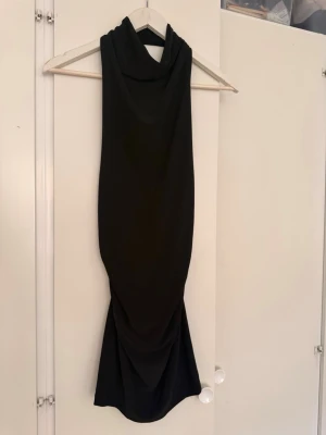 Svart lång halterneckklänning - Snygg svart långklänning med halterneck och draperad detalj framtill. Klänningen har en smal passform och är tillverkad i ett mjukt, stretchigt tyg som sitter bekvämt mot kroppen. Perfekt för dig som gillar en stilren och elegant look.