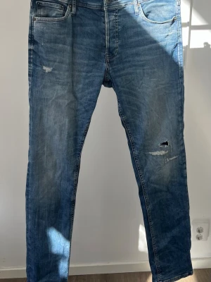 Blå slim fit jeans från Jack & Jones W33/L32 - Snygga blå slim fit jeans från Jack & Jones, modell Glenn. Jeansen har slitna detaljer och hål på ena benet för en trendig look. Klassisk femficksmodell med dragkedjegylf och normal midja. Perfekta för en avslappnad stil.