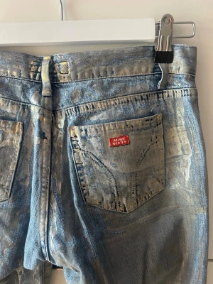 Metallicblå jeans från Miss Sixty - Säljer ett par unika metallicblå lågmidjade jeans från Miss Sixty med raka ben och klassisk femficksdesign. Jeansen har en cool glansig finish och tydliga Miss Sixty-detaljer på bakfickan. Perfekta för dig som vill sticka ut med din stil. Storlek W27, säljer då det inte passar mig längre. 