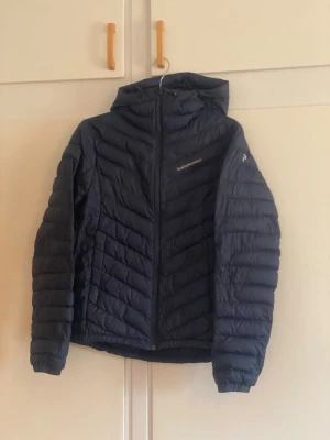Mörkblå dunjacka från Peak Performance - Snygg mörkblå dunjacka från Peak Performance med huva och dragkedja framtill. Jackan har ett quiltat mönster och är lätt vadderad för extra värme. Perfekt för kalla vinterdagar och har en sportig look.